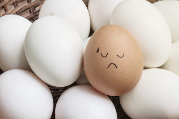 Closeup brown chicken egg with paint in tried face emotion on pile of white duck egg on wood basket background
