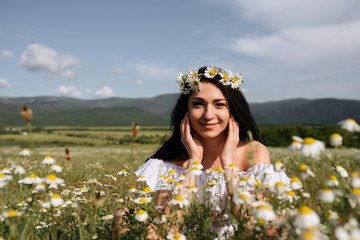 Young woman in white dress with flowers in her hands in chamomile field.