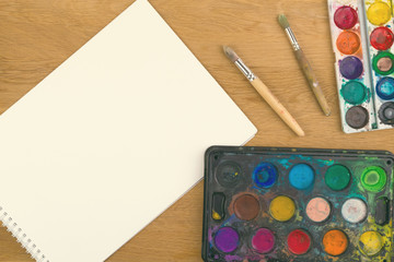 Watercolor paints, brushes for painting and blank white paper sheet on wooden background.