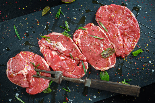 Raw Lamb leg steaks on stone chopping board with vintage fork served with sea salt and chillie flakes, rosemary and mint