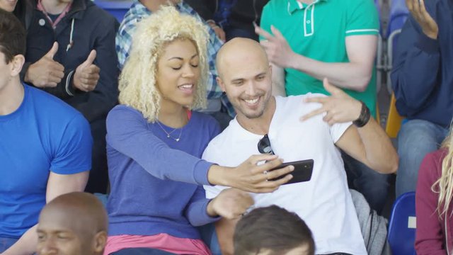  Couple sitting in the crowd at sports event pose to take a selfie with phone