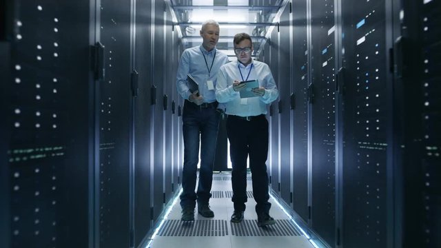 Two Server Technician Working in Data Center. One Uses Tablet Computer. They Walk Through Rows of Server Racks. Shot on RED EPIC-W 8K Helium Cinema Camera.