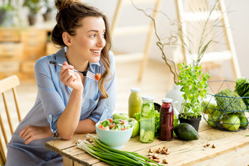 Young and happy woman eating healthy salad sitting on the table with green fresh ingredients indoors - obrazy, fototapety, plakaty