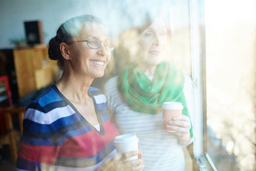 Happy senior females with hot drinks looking through window