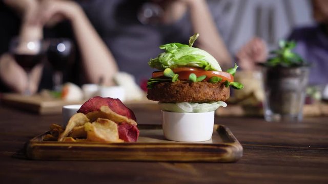 A healthy vegetable hamburger made from carrot chops, with tomato slices, lettuce leaves and soybean seedlings on onion cushion served on a wooden tray with vegetable chips and sauce