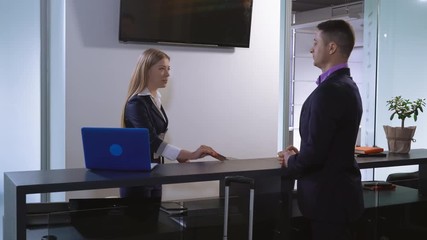 Adult man pays by credit card for room in hotel. Smiling woman using computer at working place. Worker and tourist standing near reception desk. Young receptionist talking with guest.