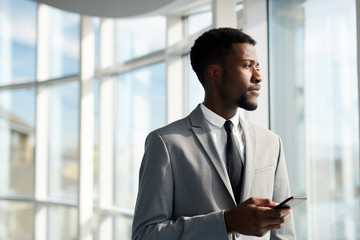 Young businessman with gadget looking through window on sunny day