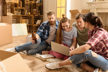 Happy young friends sitting on carpet and using laptop in new house