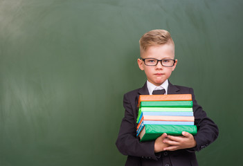 Serious boy with books standing near empty green chalkboard