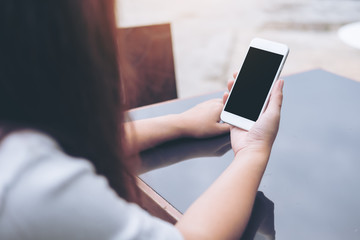 Mockup image of hands holding white mobile phone with blank black screen on vintage glass table  
