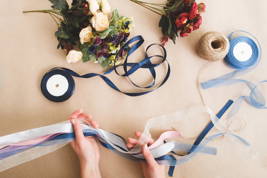 Unrecognizable person make decorations from colorful artificial flowers and ribbons on beige background, flat lay. Craftsmanship, diy art, creative hobby concept.