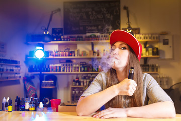 Young pretty woman in red cap smoke an electronic cigarette at the vape shop - 144756550