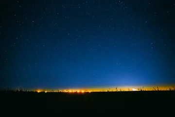 Foto auf Acrylglas Nachtsternenhimmel über dem Feld und gelbe Lichter der Stadt im Hintergrund © Grigory Bruev