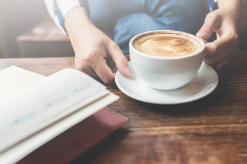 Woman holding a cup of coffee with open notebook on foreground on wooden table.