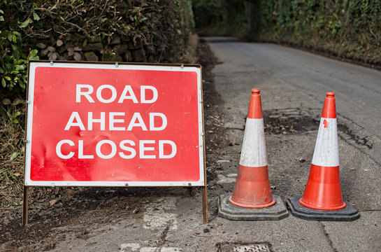 Road Ahead Closed Sign And Two Traffic Cones With Country Lane Leading Away To The Background.