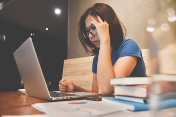 Stressed of Asian eyeglasses Woman with laptop and graph financial diagram documents on wooden table. Headache and migraine.