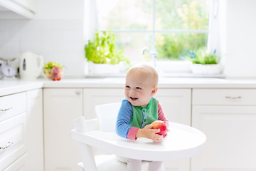 Baby boy eating apple in white kitchen at home