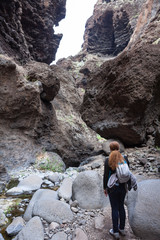 Fototapeta premium Woman hiker with backpack standing near stony pass and looking at mountain. Hiking pathway in Masca gorge. Tenerife, Canary, Spain, Europe