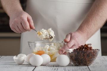 man's hands mixing cottage cheese, eggs, flour, raisins in the bowl on the table in the kitchen. Tasty, sweet meal. Healthy eating and lifestyle.