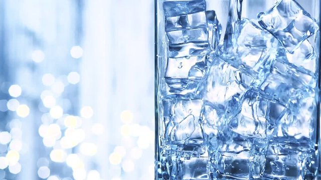 Water is pouring into a glass with ice cubes. Clear water pours ice cubes on a blue flickering background. Pure water  with Ice and bubbles in glass. Full HD  footage 1920x1080p. Slow motion 240 fps