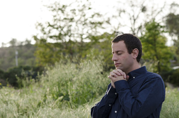 Married man praying outdoors in a grassy field with hands clasped up to his chin.