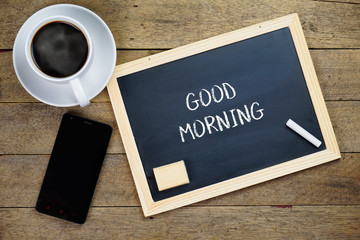 Top view of smartphone, hot coffee and chalkboard written with GOOD MORNING on wooden background.
