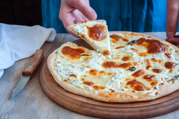 Woman's hand holding slice of homemade white pizza served on a wooden round board. Rustic style.