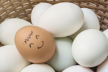 Closeup brown chicken egg with paint in smile face on pile of white duck egg on wood basket background