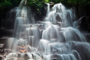 Travel in Bali jungle. Beautiful young woman sitting in yoga pose on rock under falling spring...