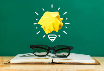 Paper yellow light bulb shine on over black glasses and notebook on wood table with green blackboard wall,Education concept