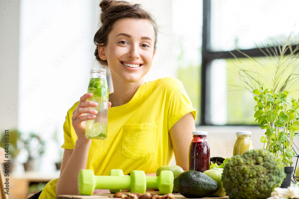 Wall mural portrait of a young sports woman in yellow t-shirt sitting indoors with healthy food and dumbbells o