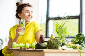 Portrait of a young sports woman in yellow t-shirt sitting indoors with healthy food and dumbbells on the table