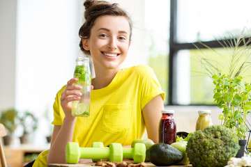 Portrait of a young sports woman in yellow t-shirt sitting indoors with healthy food and dumbbells...