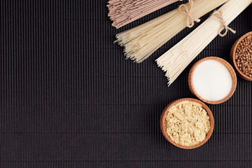 Decorative border of bundles raw noodles with ingredient in wooden bowls on black striped mat background with copy space, top view.