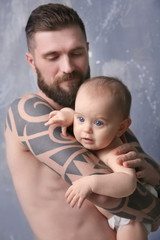Handsome tattooed young man holding cute little baby on gray background