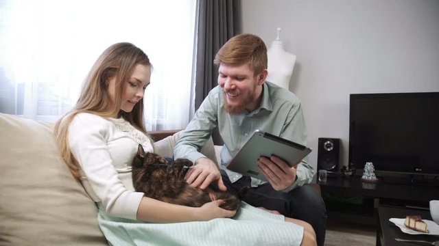 Couple is sitting on the couch sofa at home stroking a cat, and Look into the tablet and smile, close-up