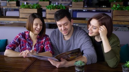 Three happy friends sitting in cafe reading menu and choosing food and beverages. Two women and one man spending lunch time together in restaurant discussing their future order. - Powered by Adobe