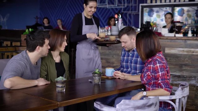 Hospitable young waitress at the cafe bringing beverages in coloured mugs to the clients. Four friends sitting at the restaurant spending their free time together and getting their order.