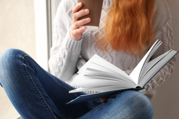Fototapeta premium Beautiful young woman drinking coffee and reading book while sitting on window sill, closeup