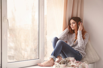 Beautiful young woman listening to music while sitting on window sill at home