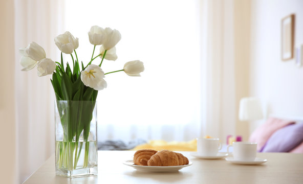 Vase with beautiful white tulips and plate with croissants on light table