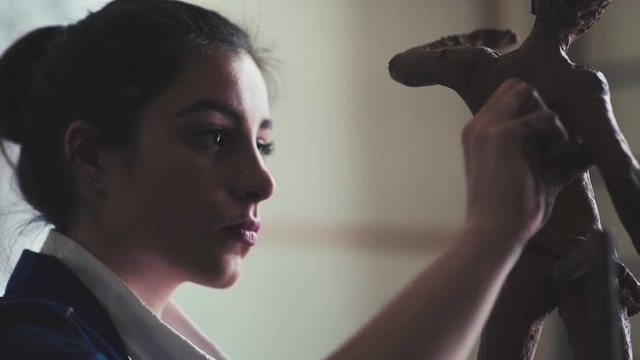 Good young woman makes a sculpture on light art studio with big window. A young student of the art academy makes a clay figure - an ancient statue in the form of a man. Clay figure is an antique man.