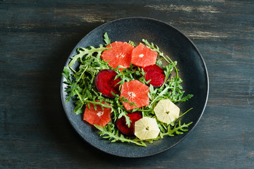Fresh mix salad of red beet, grapefruit and orange on a cushion of arugula in a black dish on a black wooden table top. View from above.