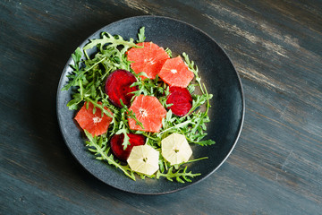Fresh mix salad of red beet, grapefruit and orange on a cushion of arugula in a black dish on a black wooden table top. View from above.