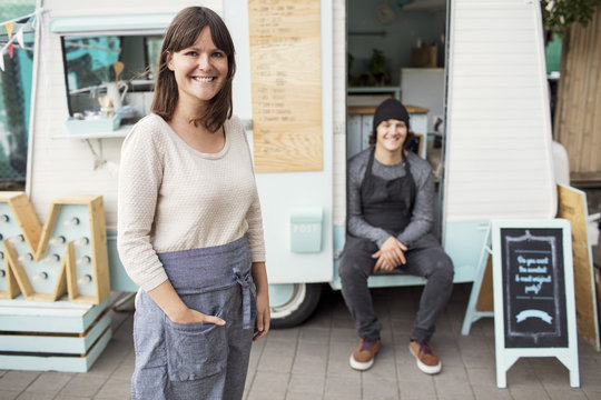 Portrait of happy female owner standing on street while colleague sitting in food truck