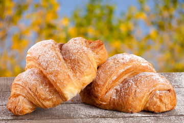 two croissant sprinkled with powdered sugar on a wooden board with a blurry garden background