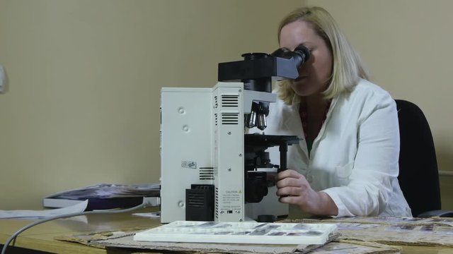 Female doctor specialist pathologist in white coat sits at a desk and using microscope to analyse a tissue samples, tumor or cancer diagnosis, laboratory analysis, portrait, interior scene.