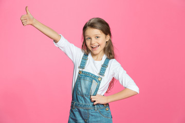 Beautiful smiling little girl in denim overalls showing thumb up on pink