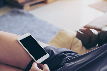 Mockup image of white mobile phone with blank white screen on woman's thigh with gray rug background in the house