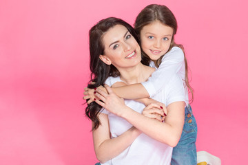 Happy beautiful mother and daughter sitting together and hugging on pink